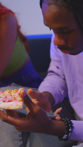 Vertical-Video-Of-Group-Of-Gen-Z-Friends-Sitting-On-Sofa-At-Home-Gaming-And-Eating-Takeaway-Pizza